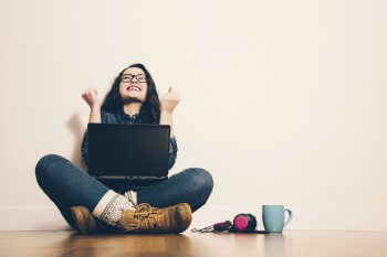 Junge Frau sitzt im Schneidersitz mit Laptop an der Wand und freut sich