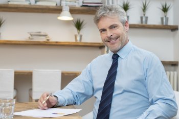 Älterer Mann sitzt mit Hemd und Krawatte und Unterlagen an einem Tisch und lächelt in die Kamera