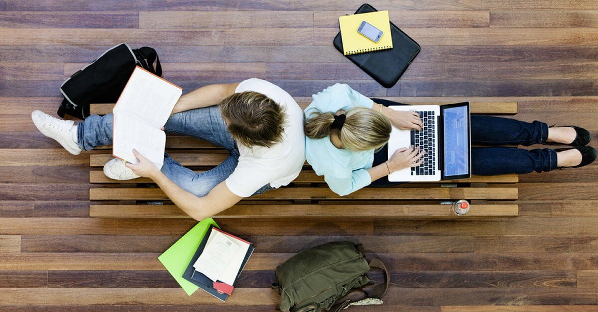 Zwei Studenten von oben, die Rücken an Rücken mit Büchern und Laptop auf dem Boden sitzen
